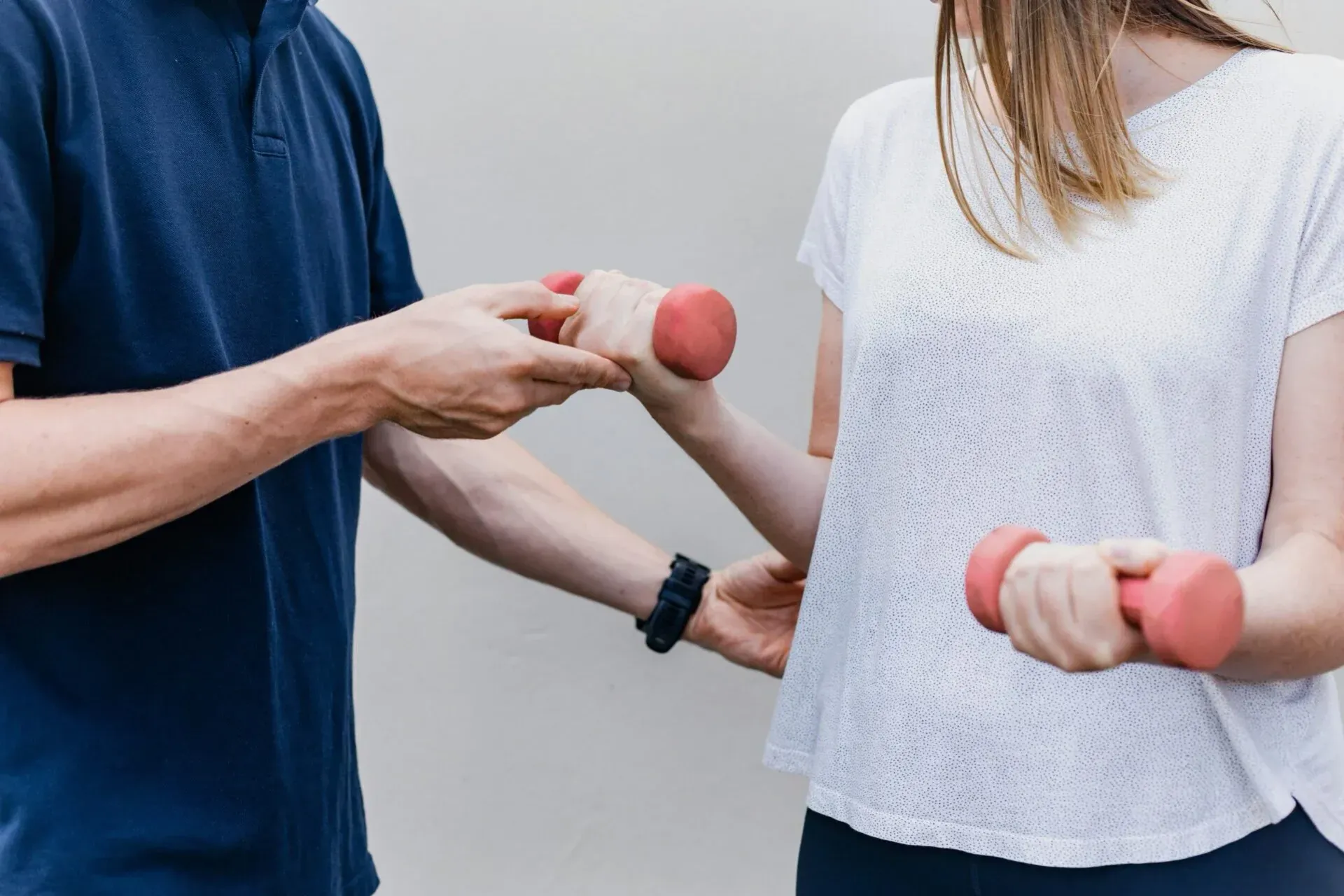 A person in a blue shirt assists another person in a white top with arm exercises using small pink dumbbells.
