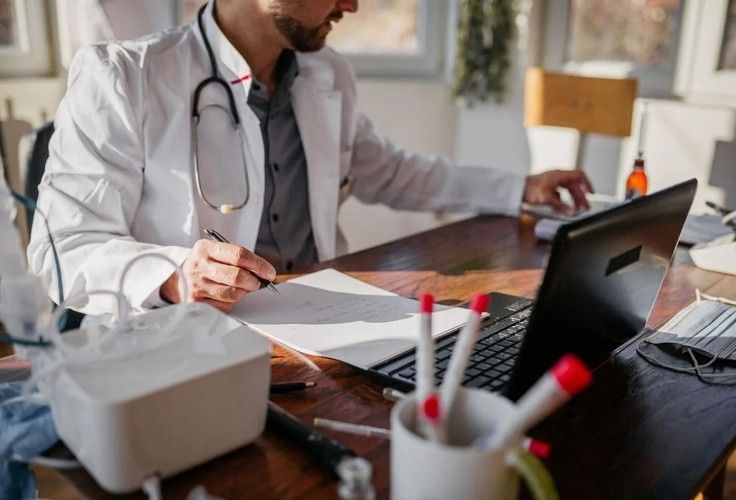 Doctor in white coat at desk with laptop, writing on paper, stethoscope, tissues, and pens.