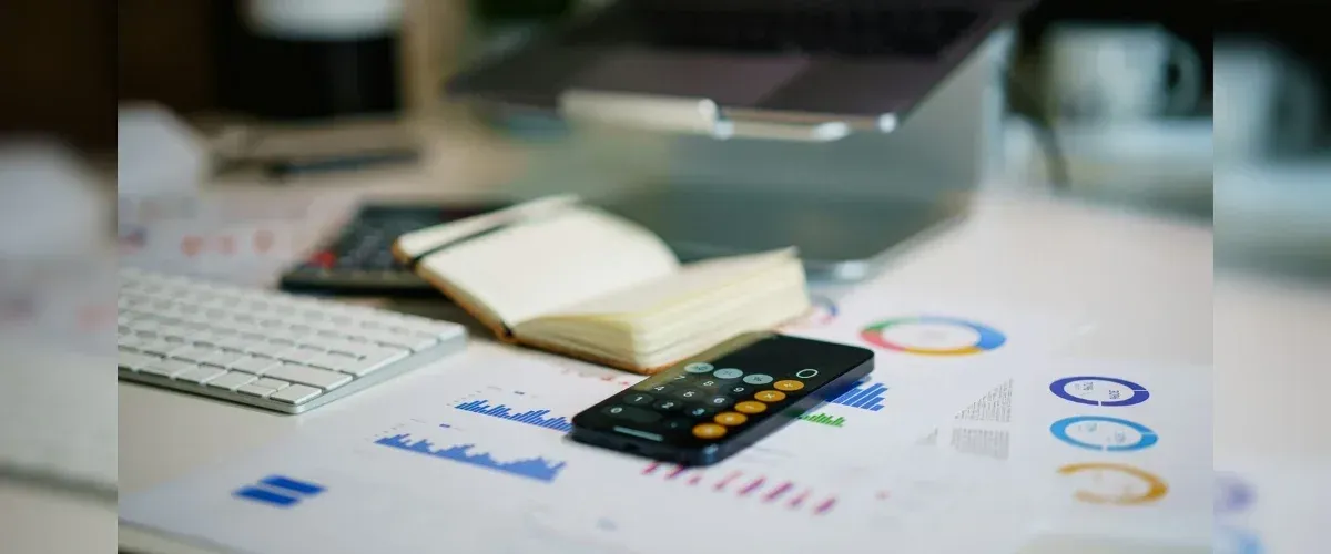 A workspace with a laptop, keyboard, open notebook, and calculator resting on charts and business data.