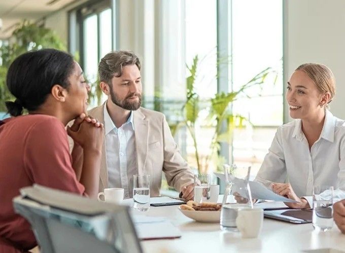 Three people in business attire at a table in a meeting. A woman is speaking with two others.