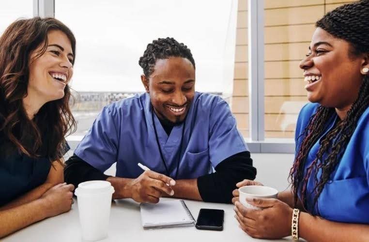Three healthcare workers laugh together at a table, likely during a break.