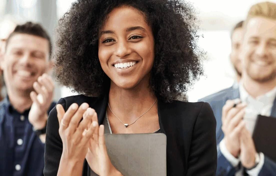 Woman in black blazer smiles and claps with colleagues in the background.