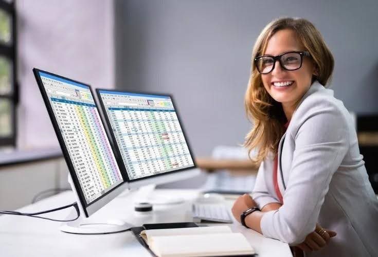 Woman with glasses smiling at a desk with two computer screens displaying spreadsheets.