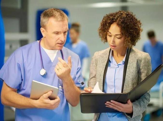A healthcare professional in scrubs consults with a woman in a blazer, both looking at a file in a hospital setting.