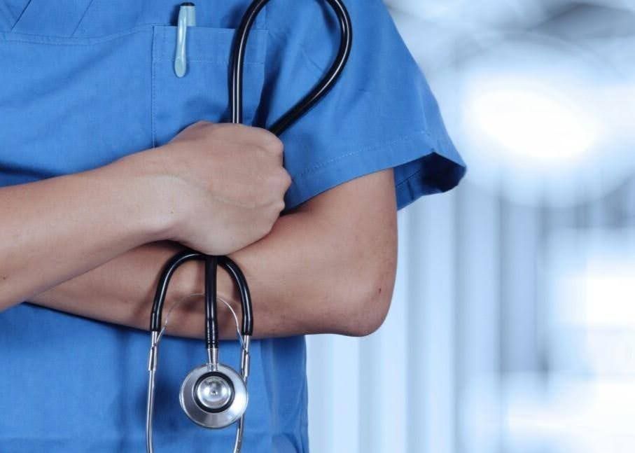 A medical professional in blue scrubs with a stethoscope around the neck and arms crossed.