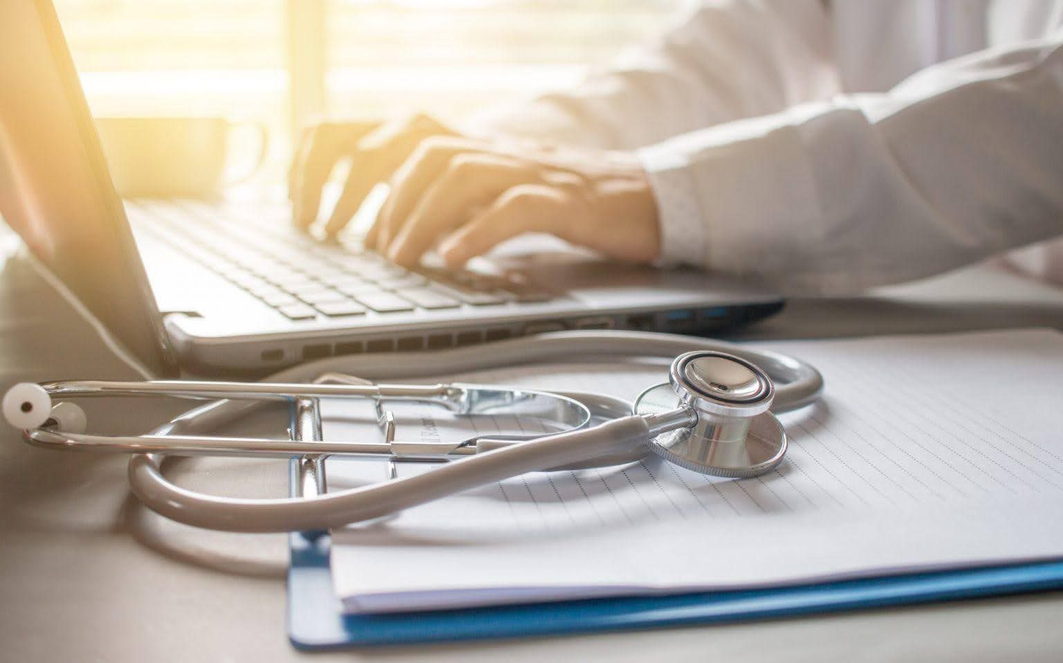 Stethoscope, laptop, and clipboard on a desk; doctor typing on laptop in bright room.