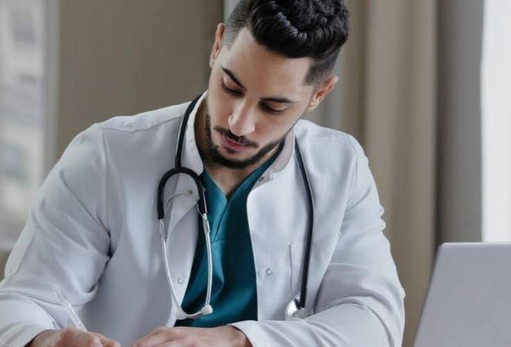 Doctor wearing a stethoscope and white coat, looking down at paperwork, near a laptop.