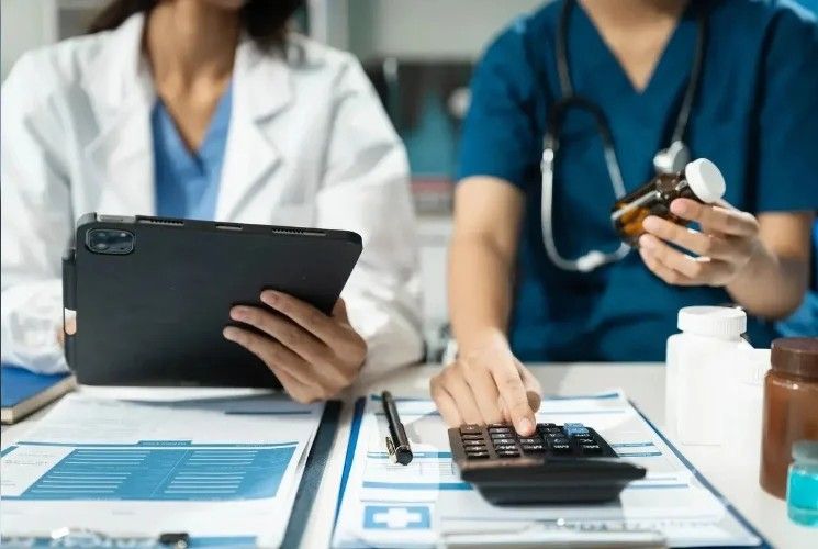Two medical professionals reviewing documents, one using a tablet, the other with a calculator and medication bottles.