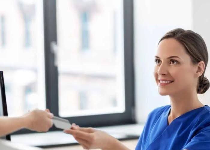 Nurse in blue scrubs smiling while receiving a card from a person off-screen in a bright office.