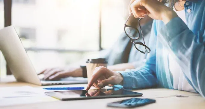 Person using a tablet and another working on a laptop at a bright office desk; eyeglasses, coffee cup, and phone visible.