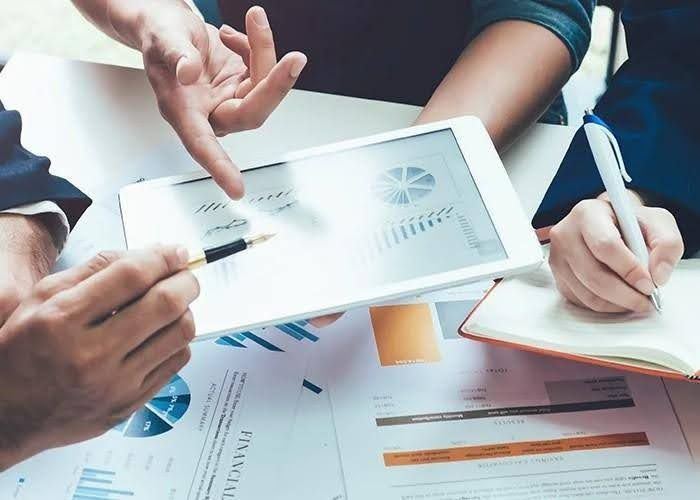 People discussing charts on a tablet, pointing with a pen, papers, and notebook on the table.