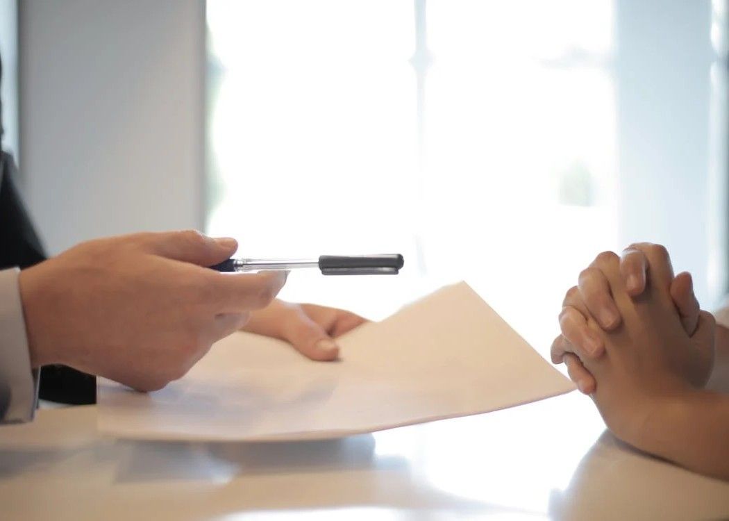 A person hands a pen across a table to another person over a blank document, suggesting a professional sign-off.