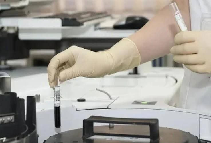 Person in gloves holding vials in a laboratory setting, with machinery and other equipment visible.