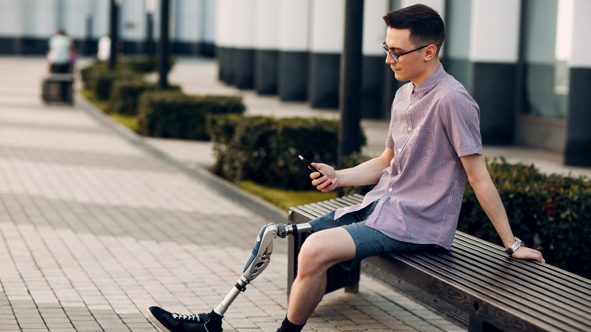 Man with a prosthetic leg sits on a bench, looking at his phone in an outdoor setting.