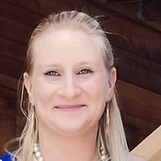 Blonde woman with fair skin, smiling; outdoors with wooden background, wearing earrings.