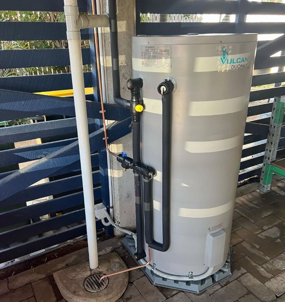 A Vulcan water heater outdoors, with pipes and fittings. It is next to a fence and on a concrete pad. — Off-Tap Plumbing NQ In Currajong, QLD
