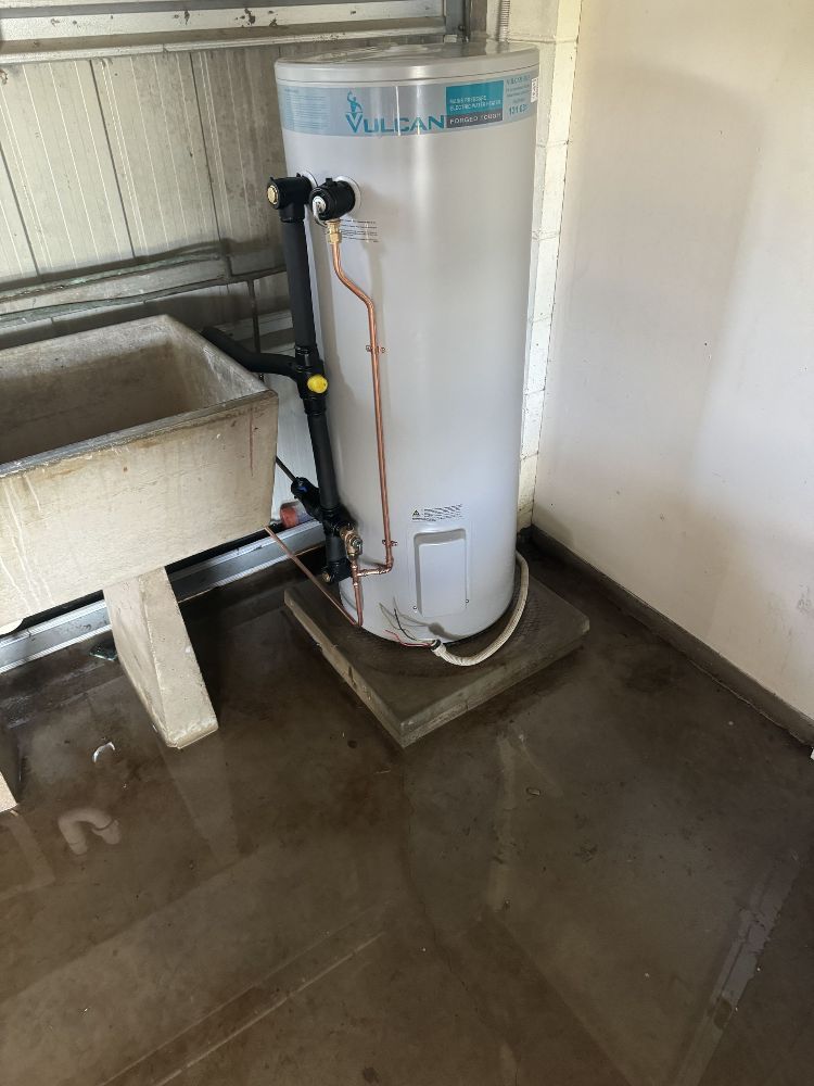 Water heater in a garage, standing on a concrete pad in a pool of water. A utility sink is to the left.— Off-Tap Plumbing NQ In Currajong, QLD