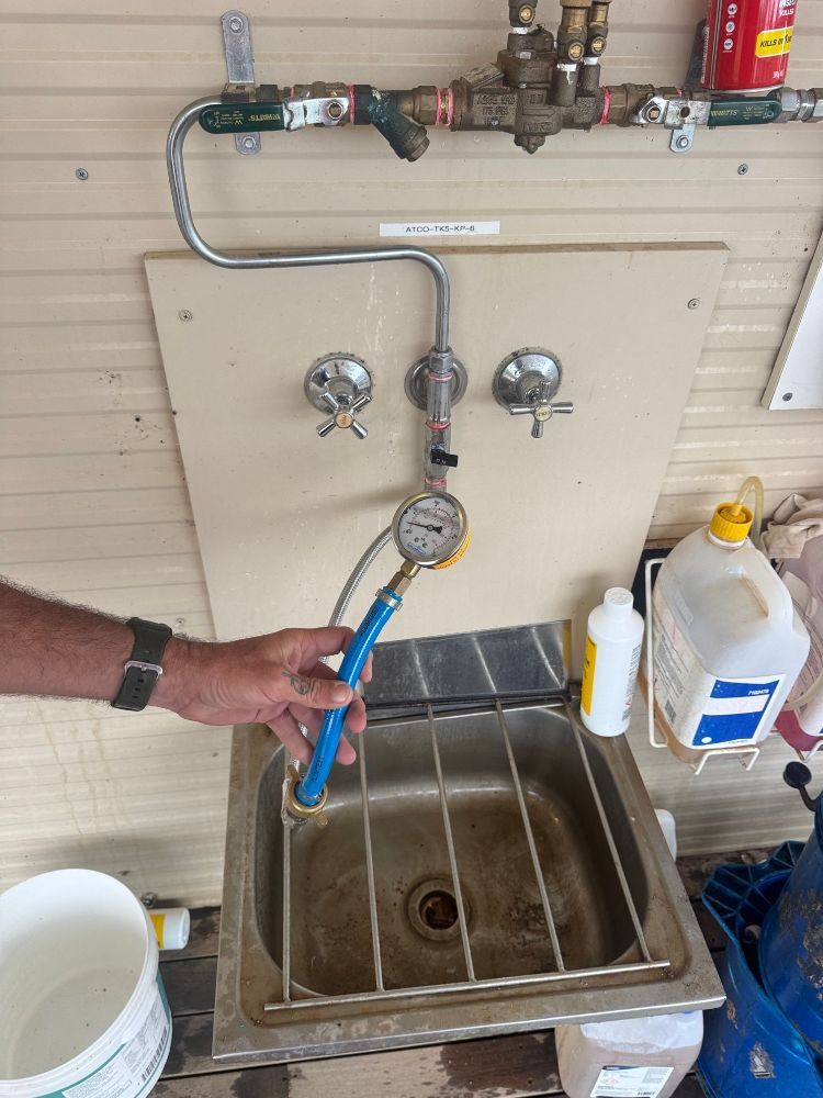 A person holds a blue hose connected to a pressure gauge above a dirty stainless steel sink. Plumbing fixtures are mounted on a wooden wall.— Off-Tap Plumbing NQ In Currajong, QLD