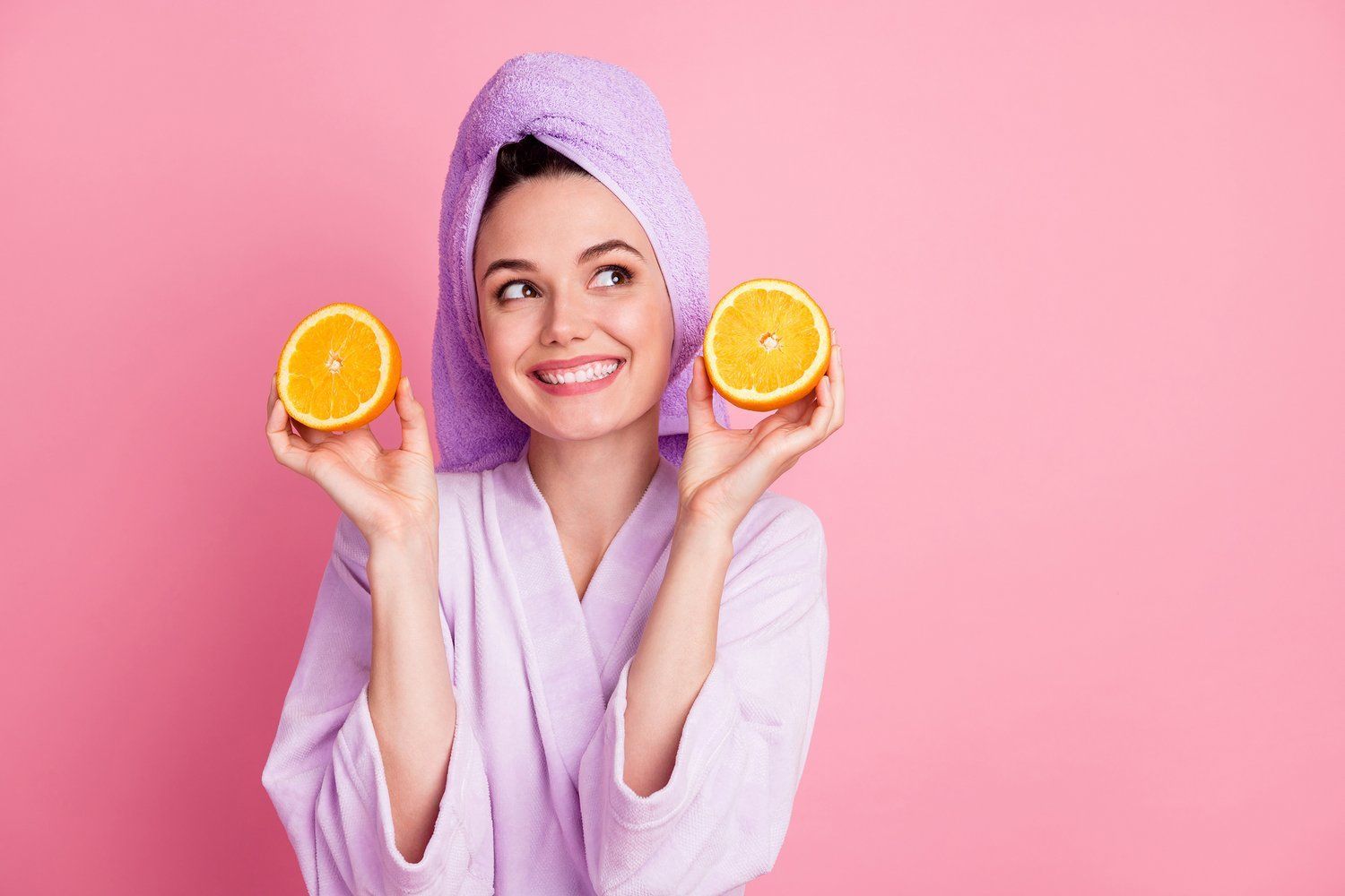 A woman with a towel wrapped around her head is holding two slices of orange.