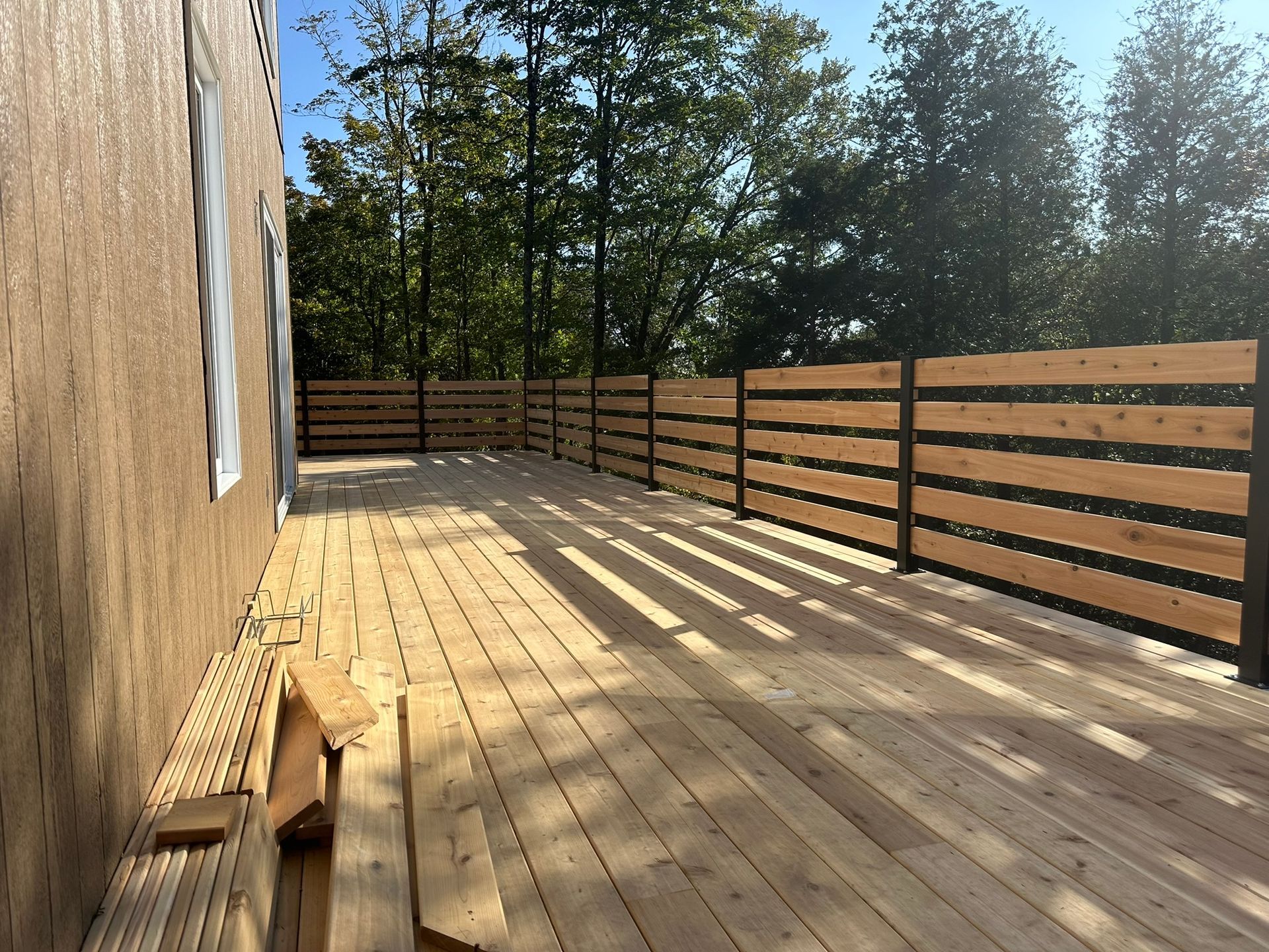 Terrasse en bois avec une clôture à lattes horizontales jouxtant un bâtiment. Arbres en arrière-plan.