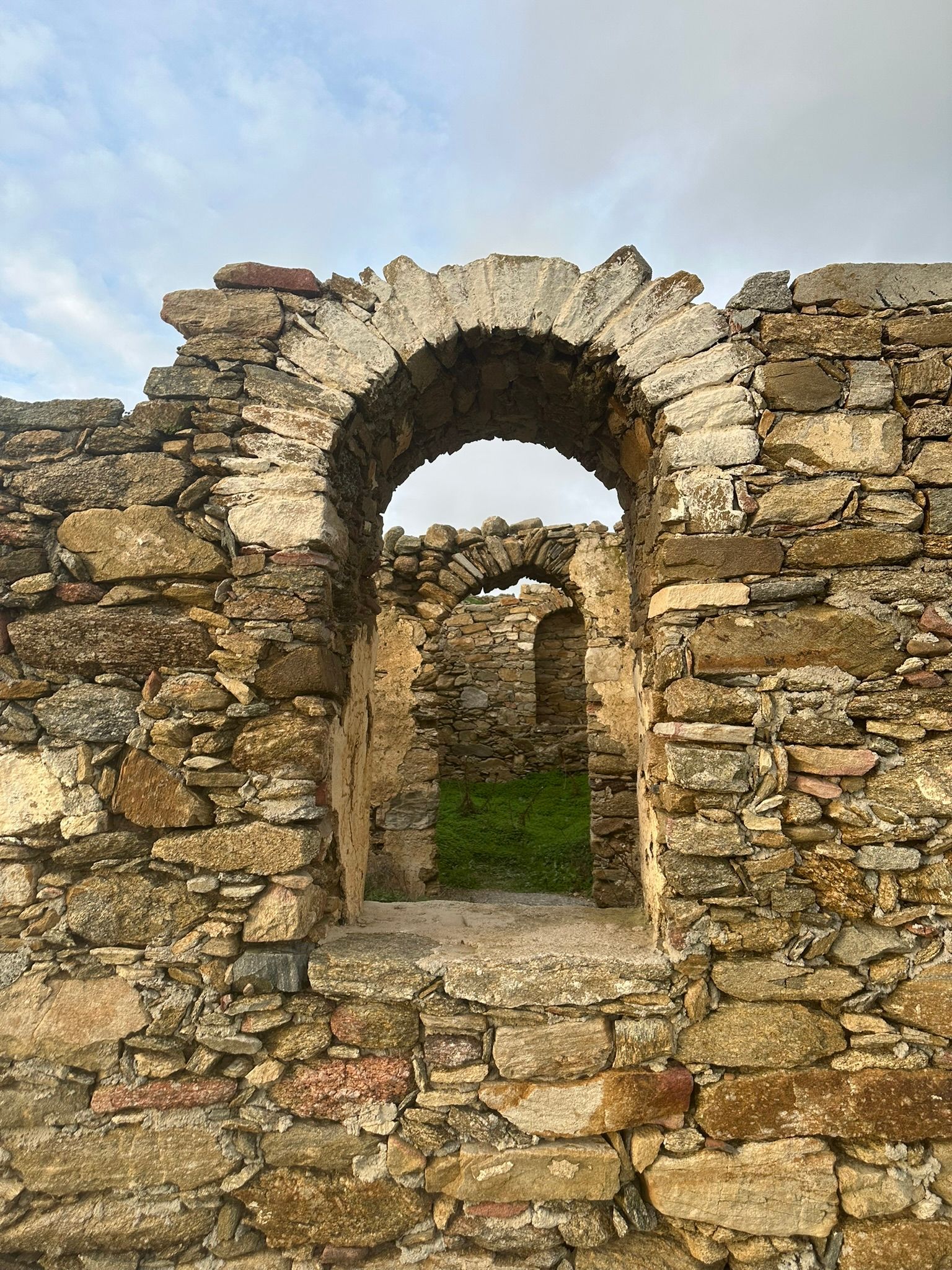 Ruines de pierre avec une porte cintrée donnant sur d'autres ruines, ciel nuageux.