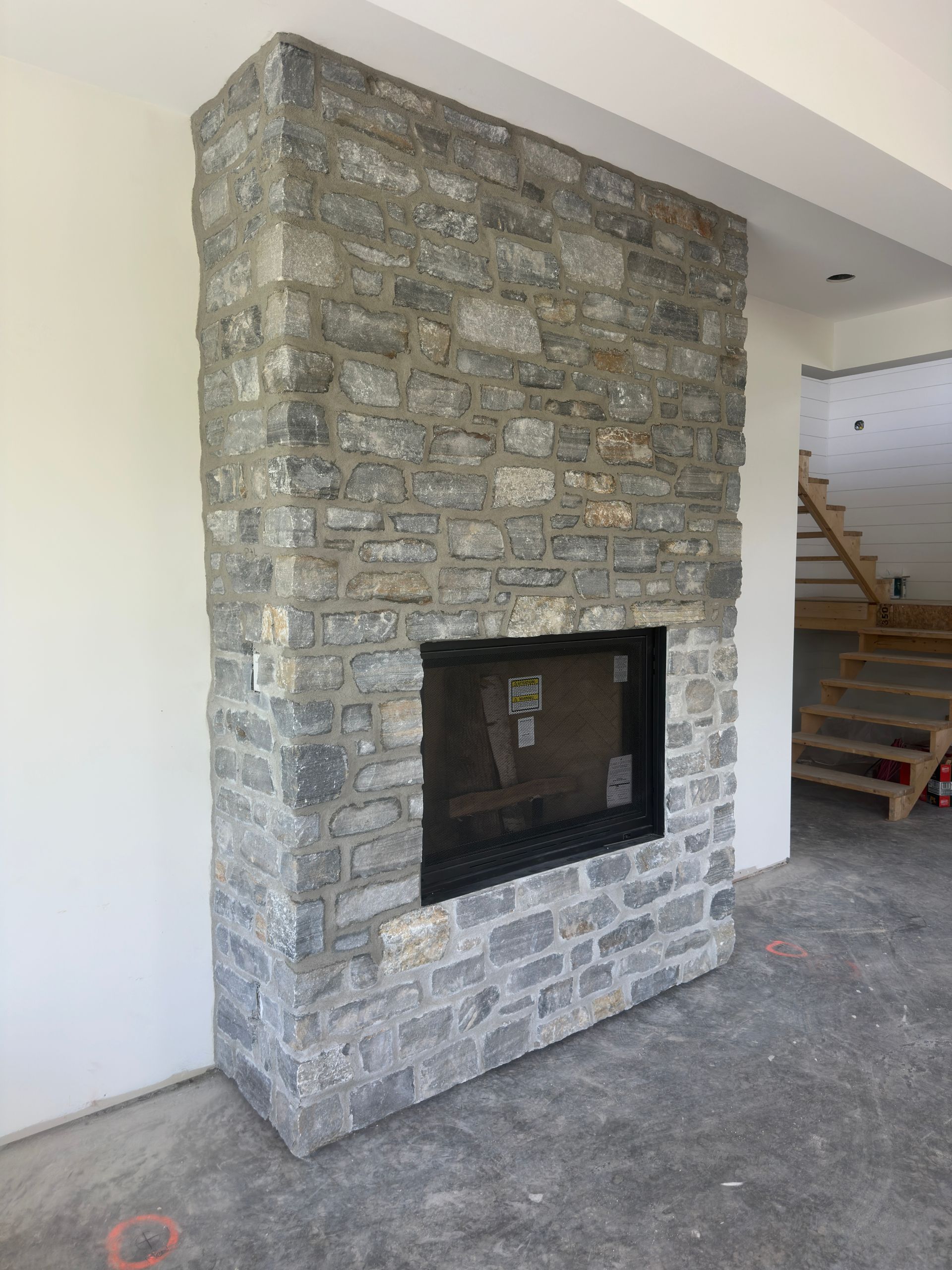 Cheminée en pierre adossée à un mur blanc. Foyer à cadre noir au centre.