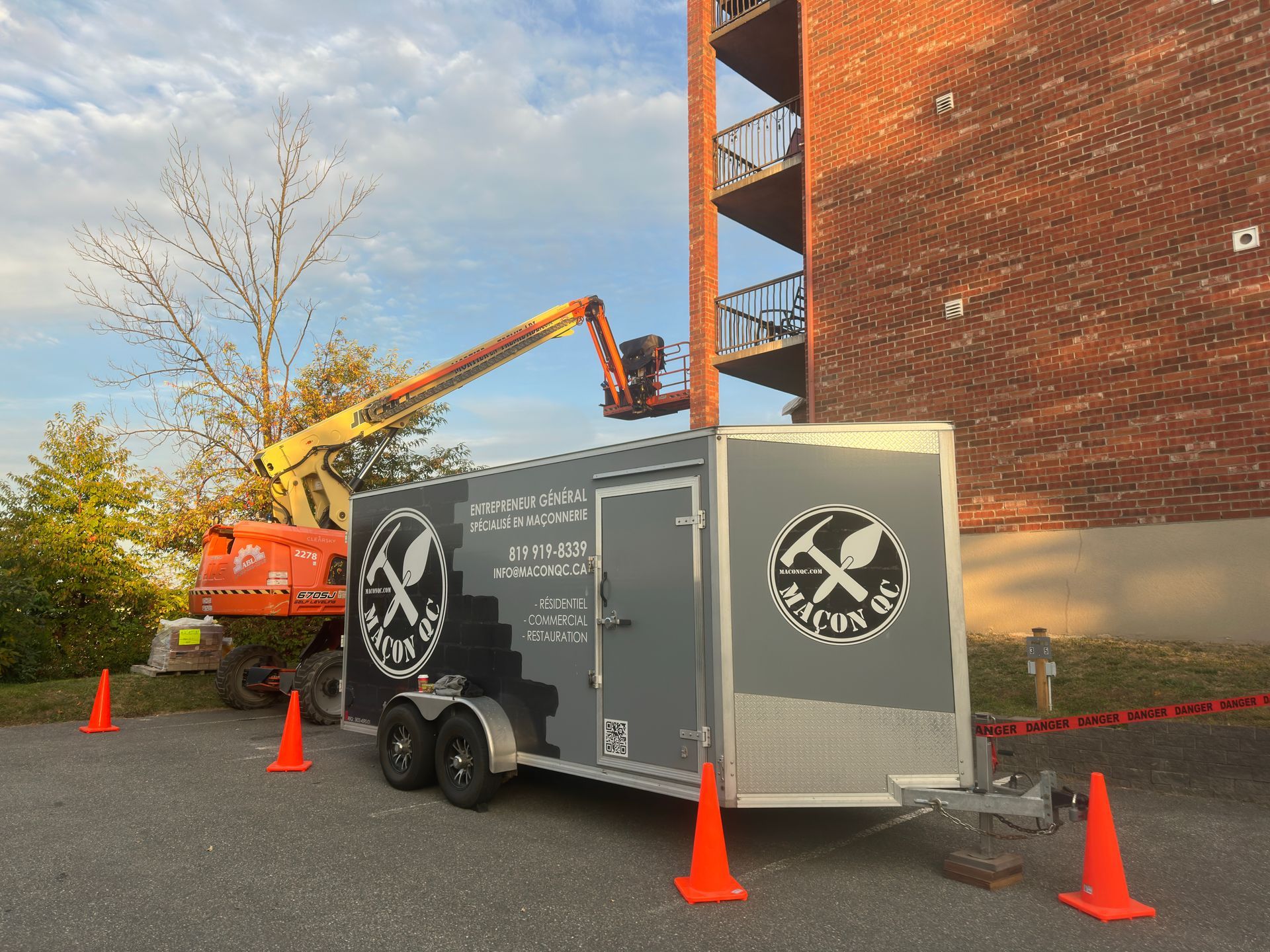 Une remorque de chantier avec logo et nacelle orange près d'un bâtiment en briques, avec des cônes de signalisation et du ruban de signalisation.