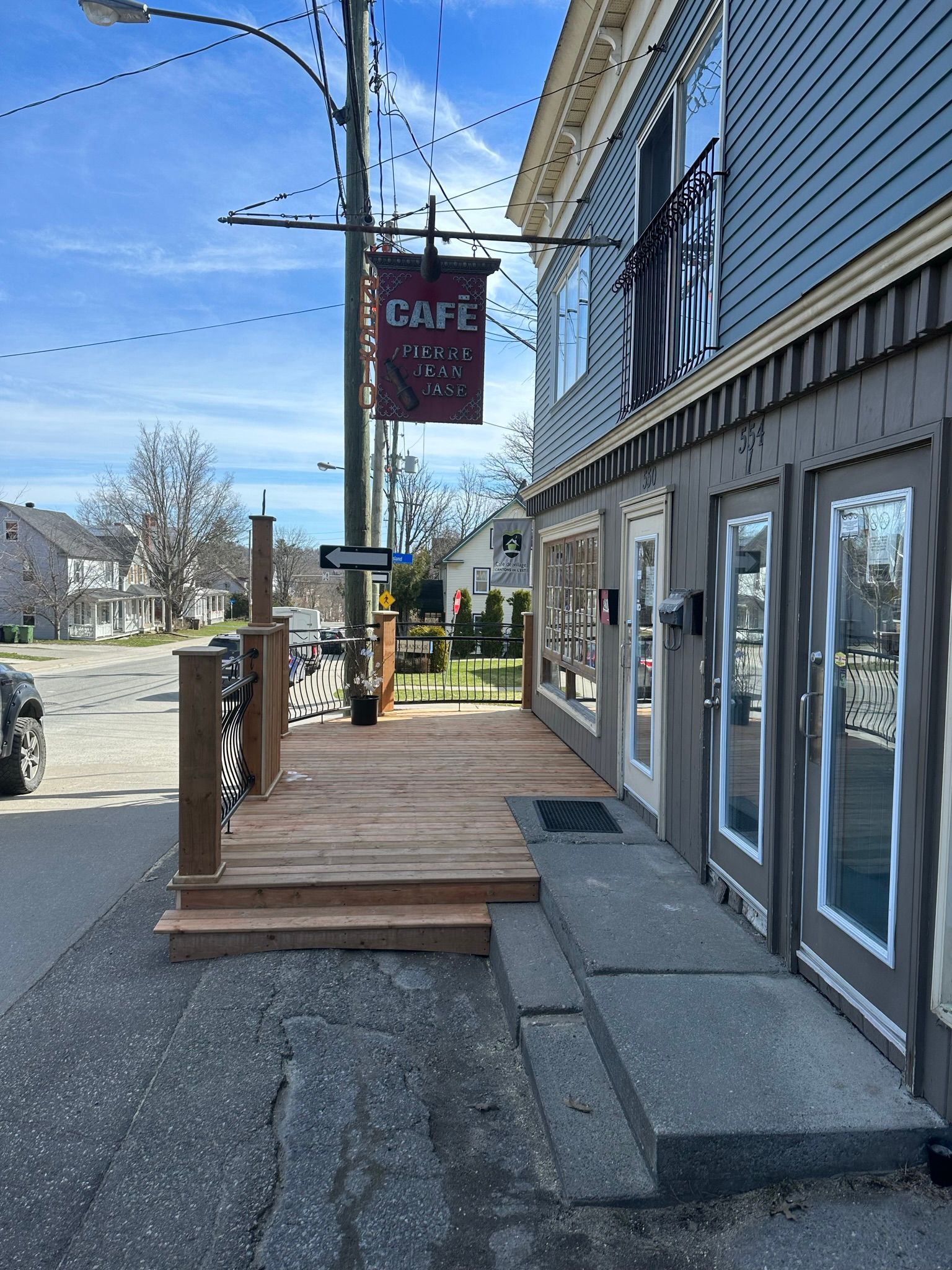 Façade de café avec terrasse en bois, rampe et enseigne suspendue par une journée ensoleillée.