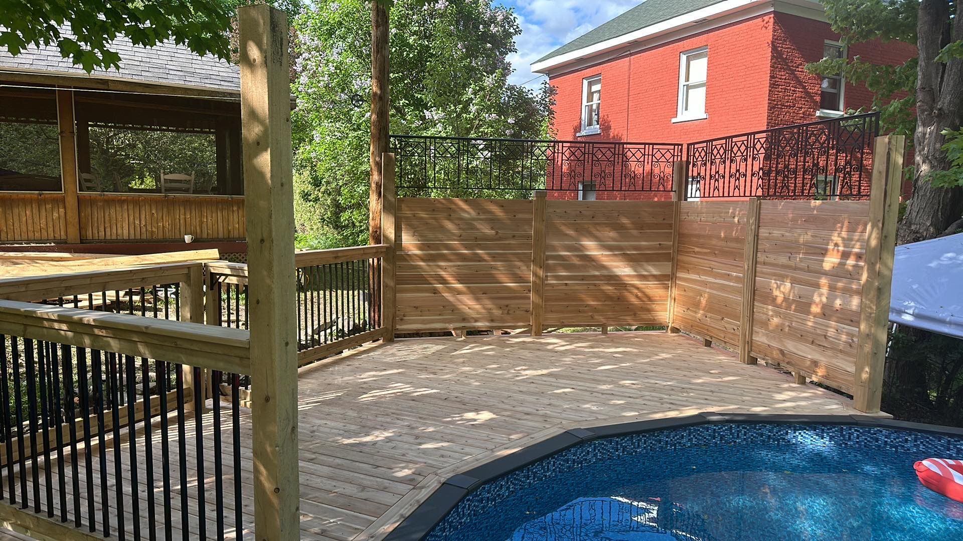 Terrasse en bois avec piscine hors sol, gazebo et maison rouge en arrière-plan. Journée ensoleillée.