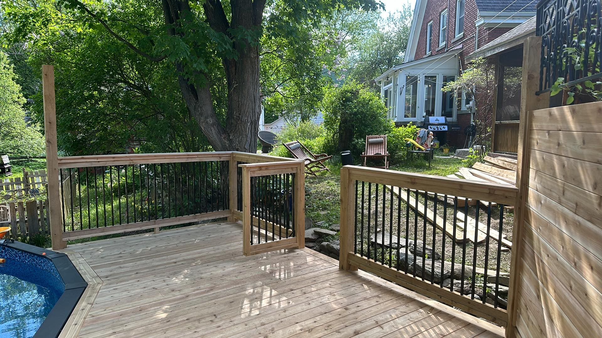 Terrasse en bois avec balustrade noire et portail près d'une piscine et d'une maison avec un jardin verdoyant.