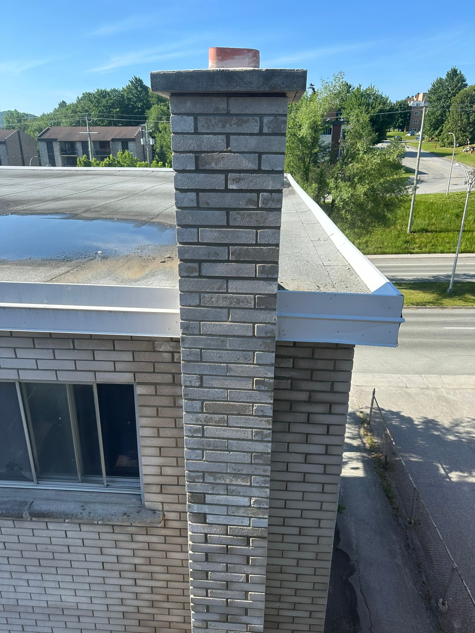Cheminée en briques sur un toit plat, briques grises, ciel bleu, bâtiments en arrière-plan.