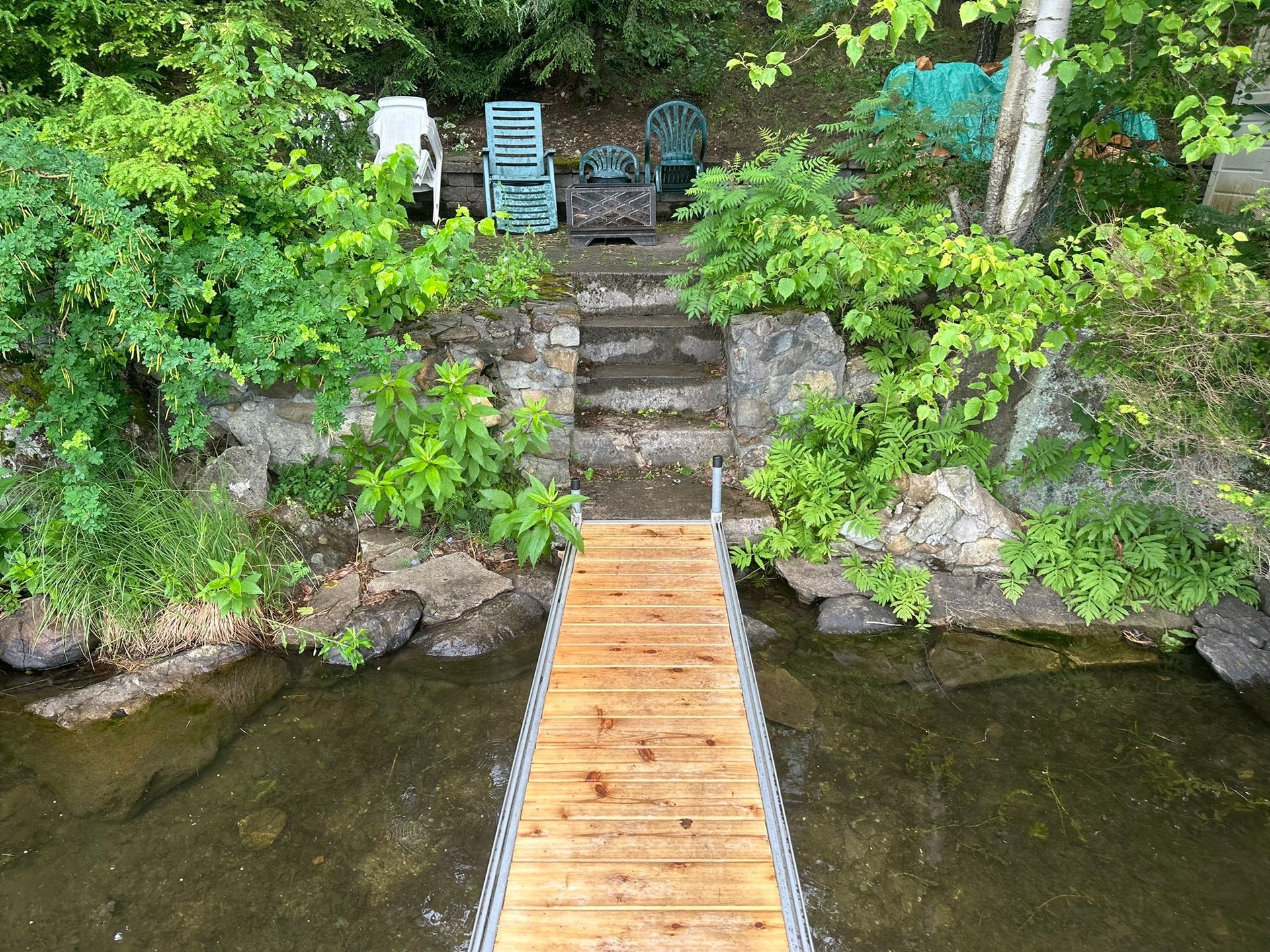 Un quai en bois mène à des marches en pierre et à des chaises sur une berge herbeuse, entourée de verdure.