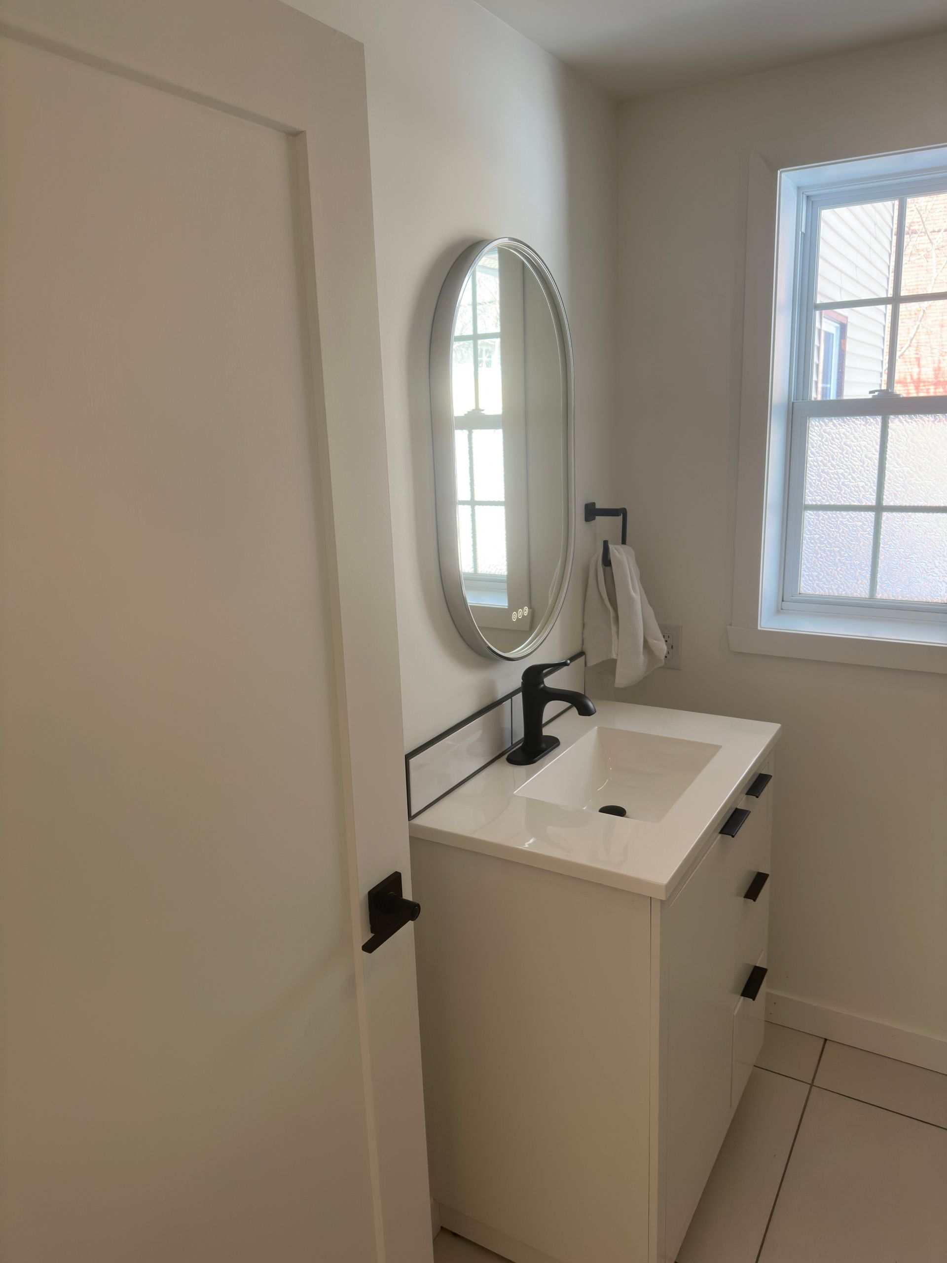 Petite salle de bains blanche avec lavabo, miroir et fenêtre. Robinetterie et poignées de porte noires.
