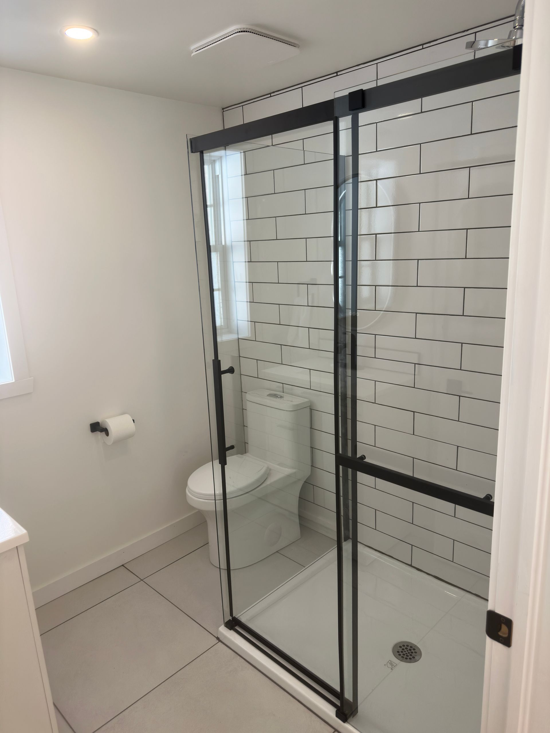 Salle de bains avec douche carrelée blanche, porte vitrée à cadre noir et toilettes blanches.