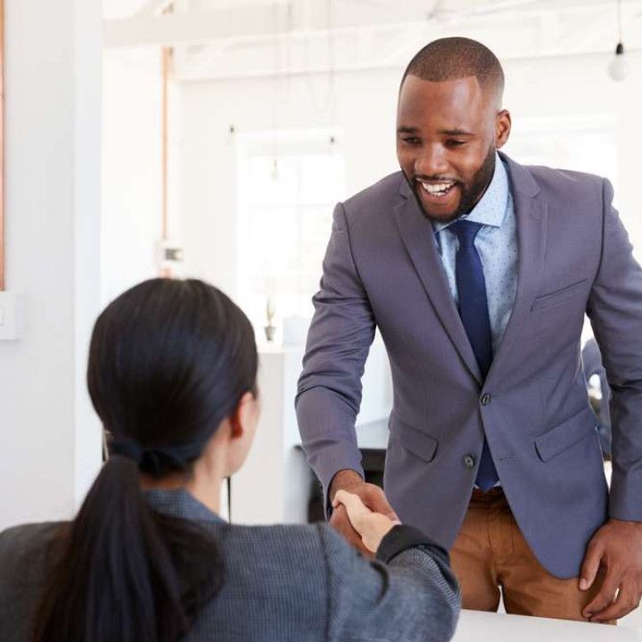 A smiling Black man in a suit shakes hands with a woman in a grey suit in an office.