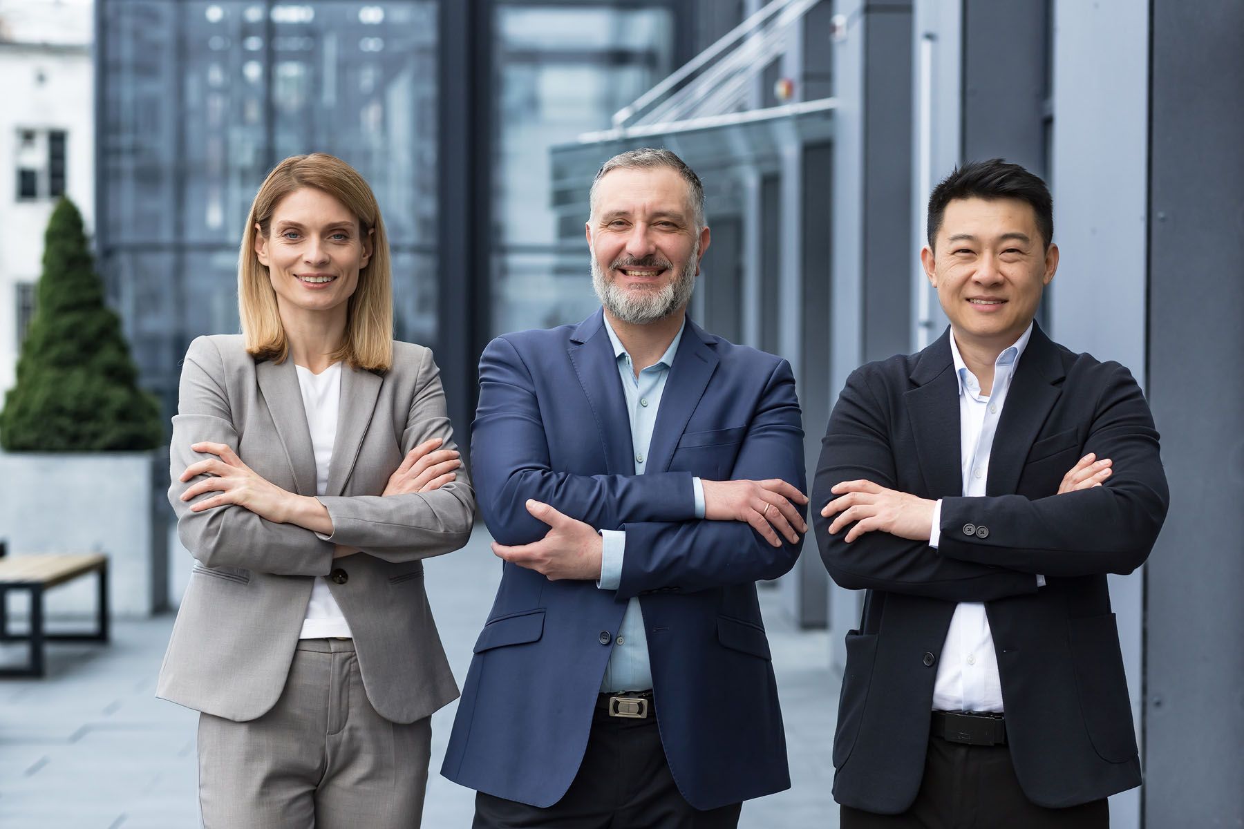 Three diverse business professionals, arms crossed, smiling confidently outside a modern building.