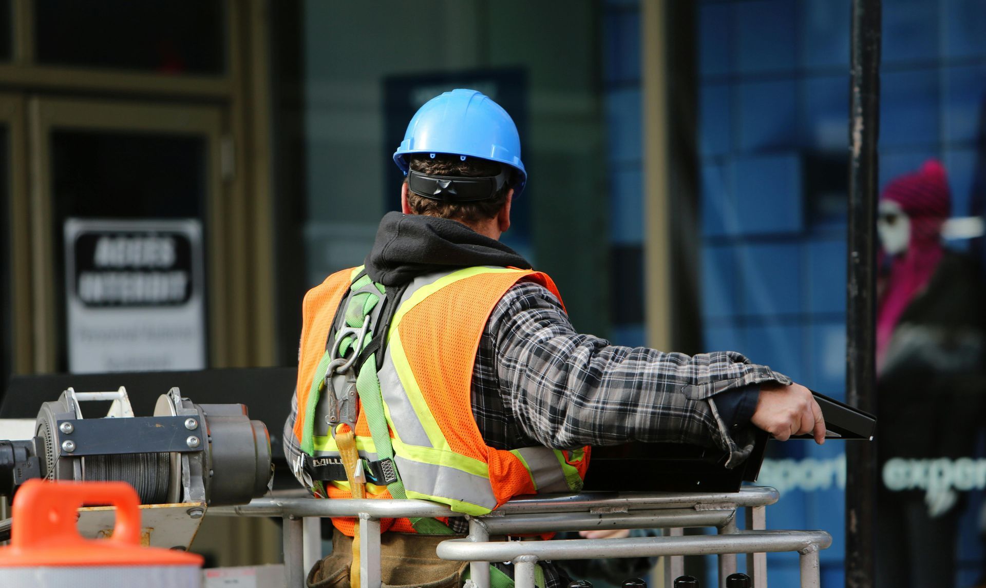 Construction worker in blue helmet, orange vest, holding a device, standing outdoors.