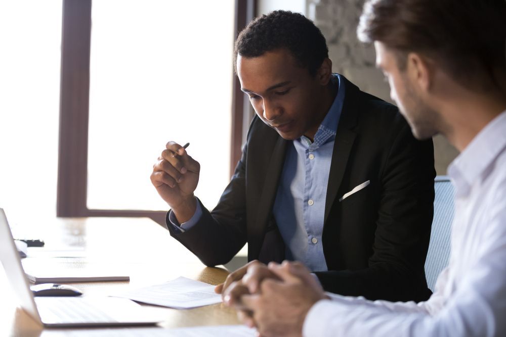 Two men reviewing documents at a table in an office setting. The man on the left is Black and holding a pen.