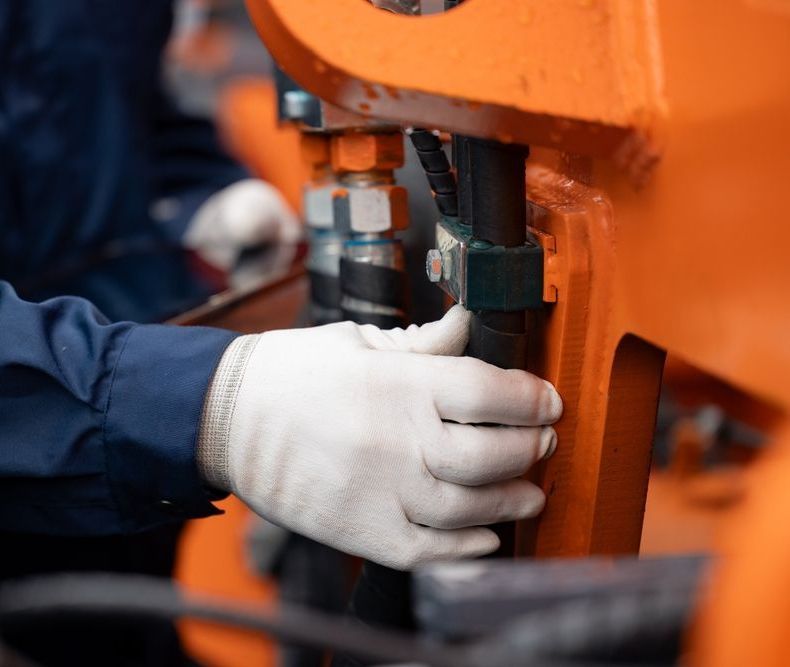 Mechanic wearing gloves, adjusting hydraulic hoses on orange industrial equipment.