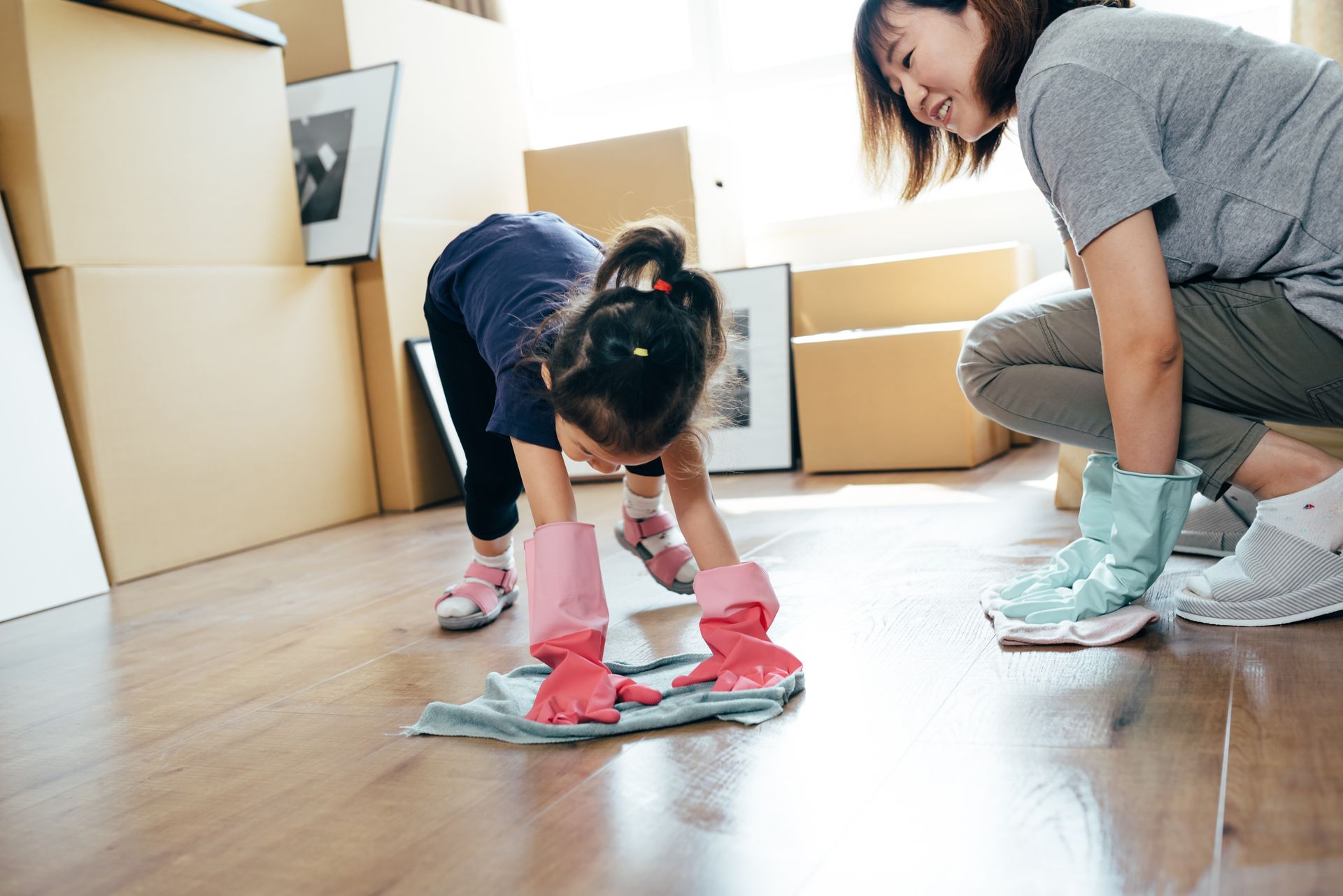 Child wearing gloves cleaning wooden floor with cloth while adult assists during home cleaning.