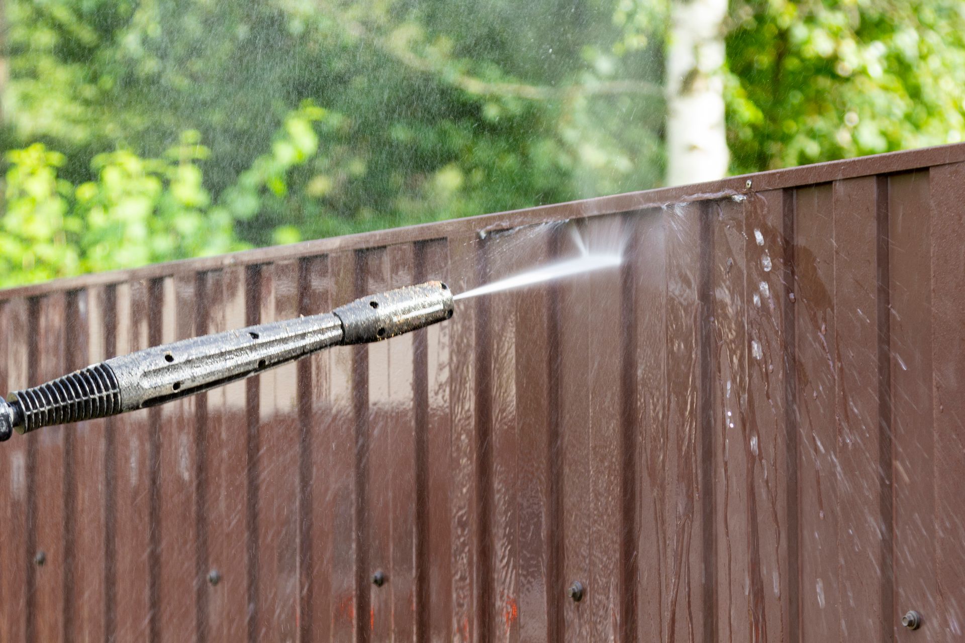 Pressure washer cleans a brown fence with water, with a blurry background with leaves and trees.