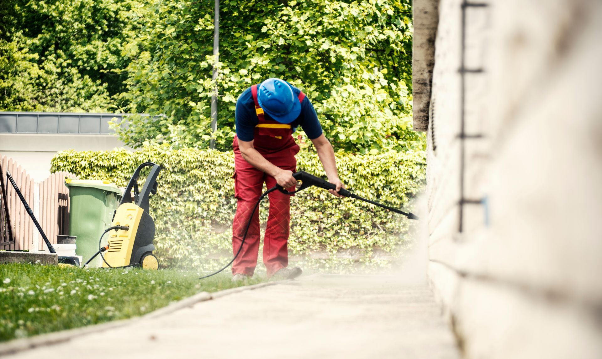 A man in red and blue work clothes is pressure washing the yard to remove dirt and debris.
