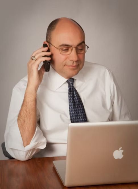 Man in white shirt on a phone call, looking at laptop.