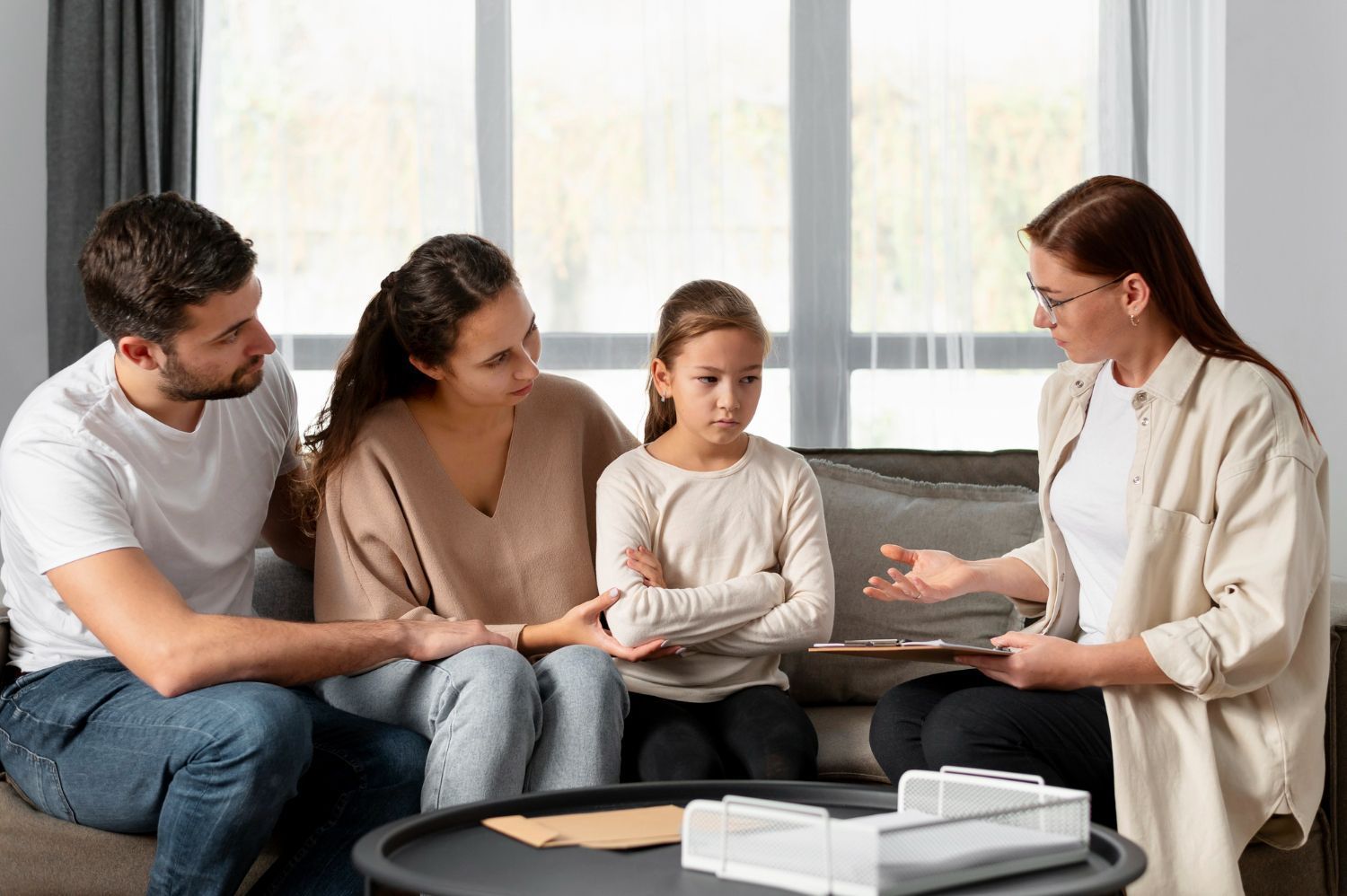 Family and therapist in a counseling session. They are seated on a couch indoors; the therapist holds a clipboard.
