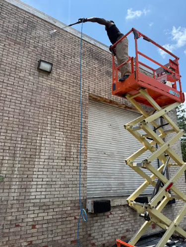 Man on an orange lift pressure washing a brick building, blue sky in background.