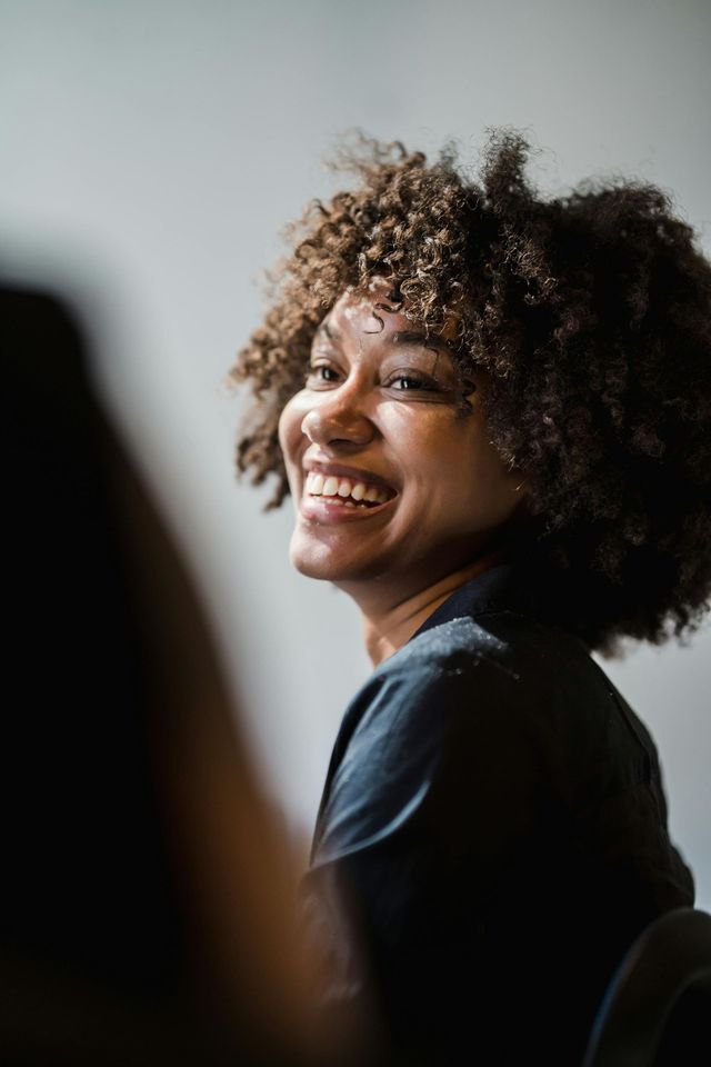 Smiling person with curly hair in a dark shirt, turned toward the camera against a soft gray background