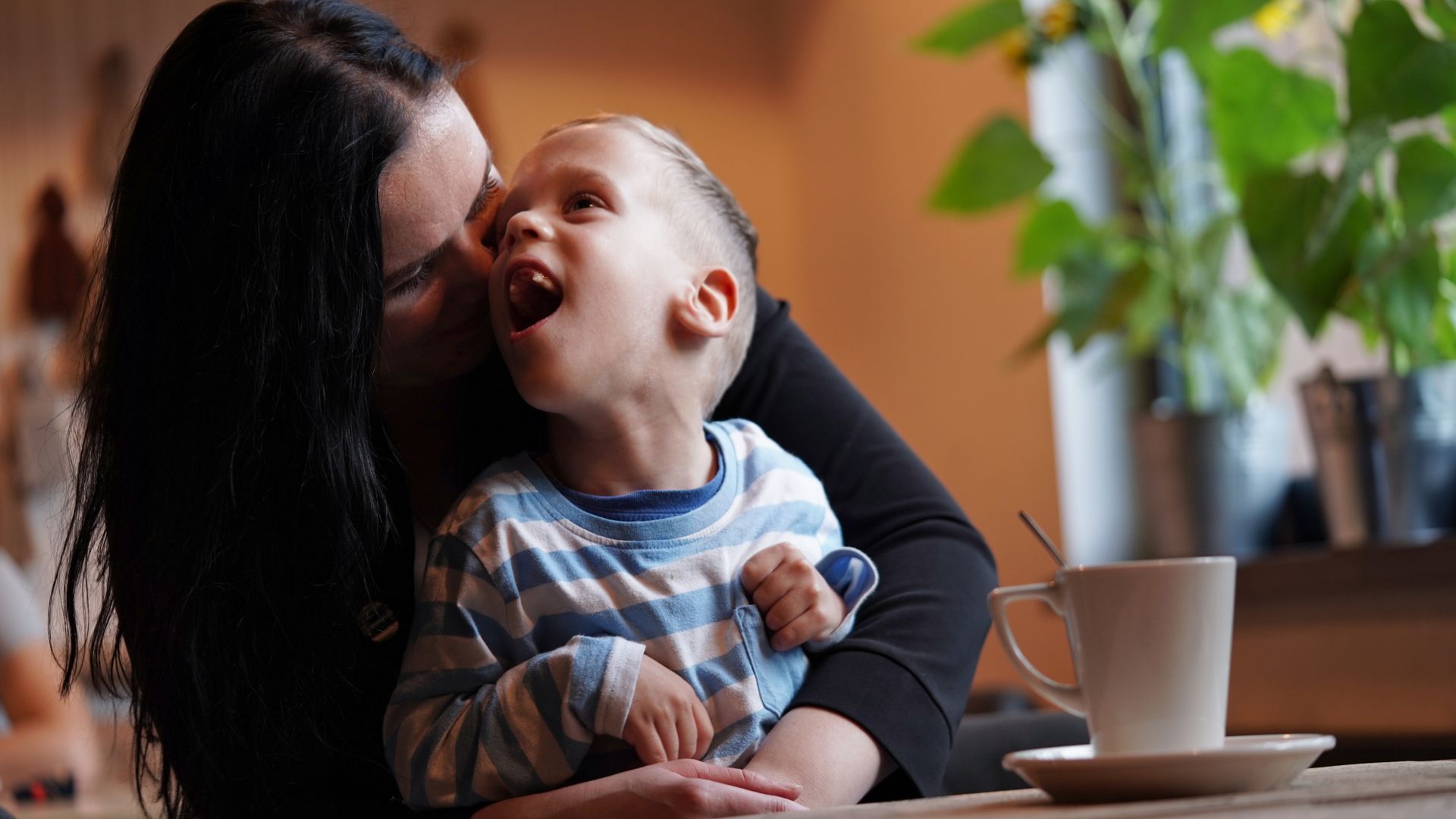 A woman is kissing a baby on the cheek while sitting at a table with a cup of coffee.
