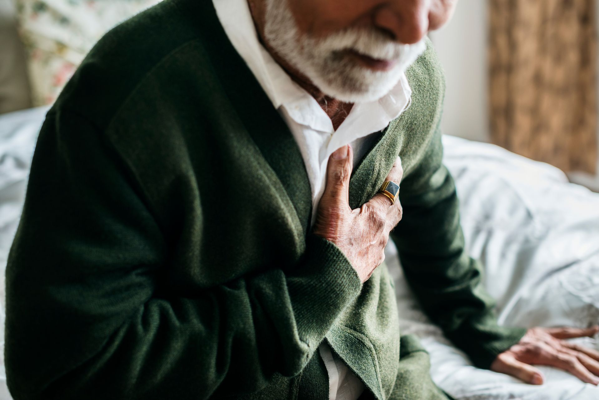 An elderly man is sitting on a bed with his hand on his chest.