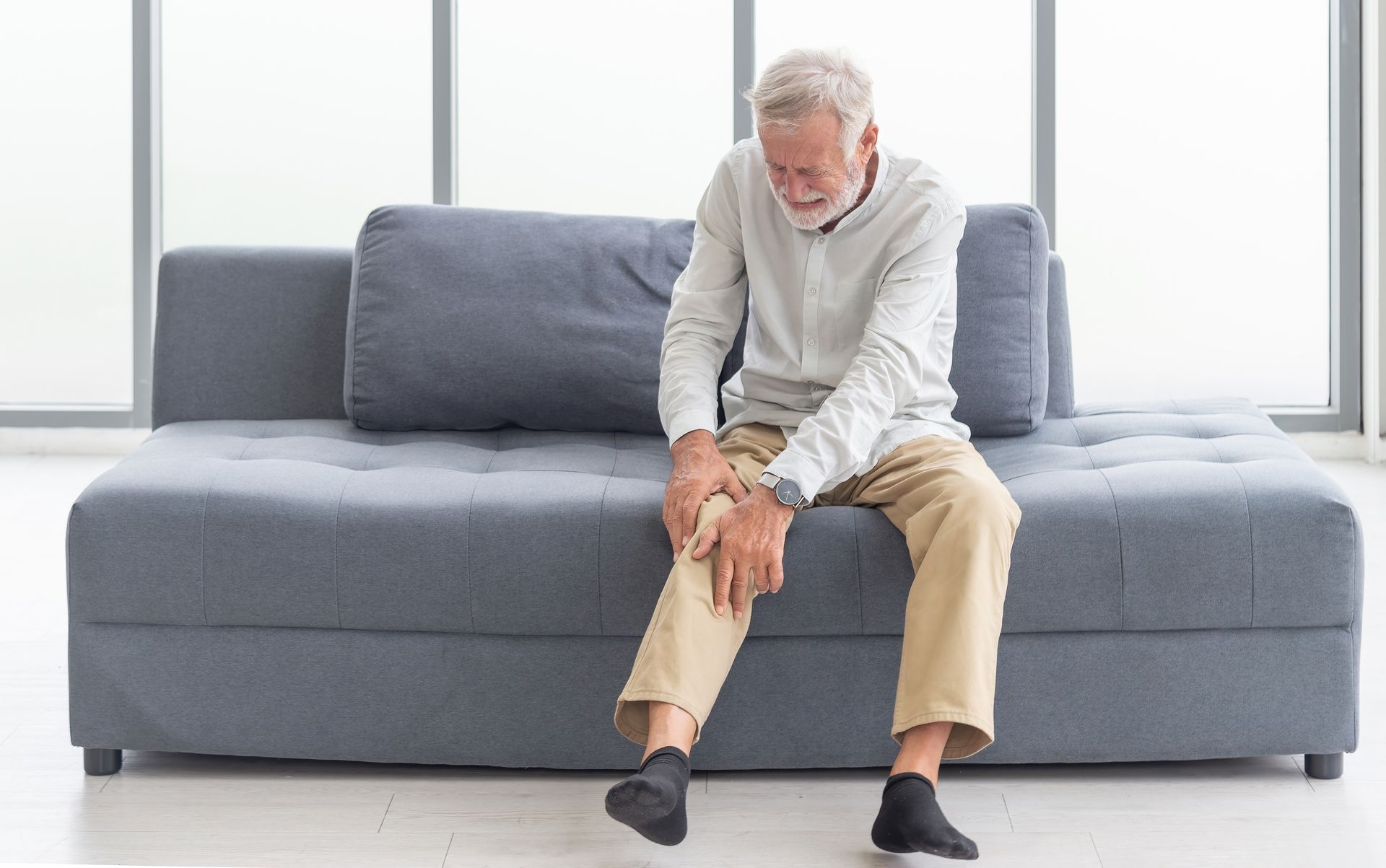 An elderly man is sitting on a couch holding his knee in pain.