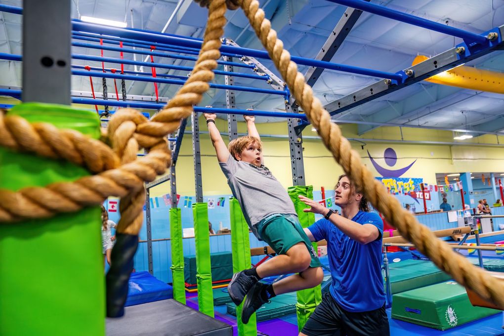 A man is helping a young boy climb a rope in a gym.